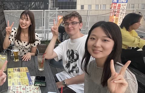 Three people smile and make peace signs while seated at a table on an outdoor city patio.