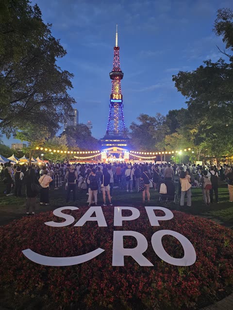 A crowd gathers in a park at dusk before the illuminated Sapporo TV Tower, with a large SAPPORO sign in the foreground.