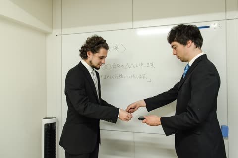 Two men in suits exchange business cards in front of a whiteboard with Japanese writing.