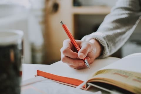 Person's hand writing with a red pen in an open notebook.