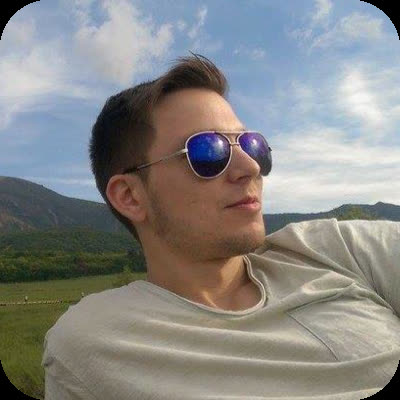 A man in reflective aviator sunglasses and a t-shirt looks to the side in a mountainous setting.