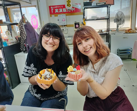 Two smiling women hold up small, decorative replica cakes with animal and fruit figures inside a workshop.